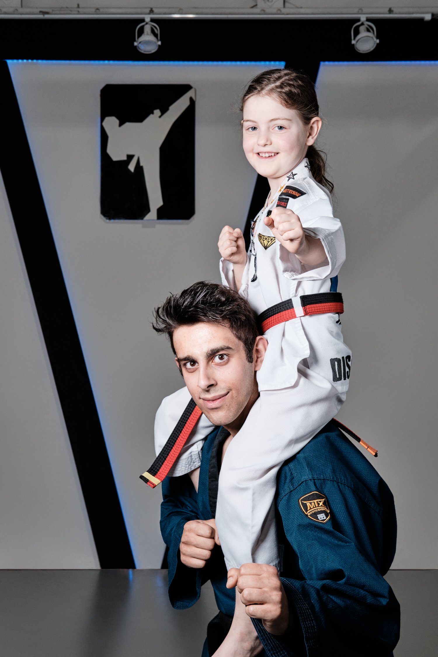 A smiling young taekwondo student sits on the instructor's shoulders.