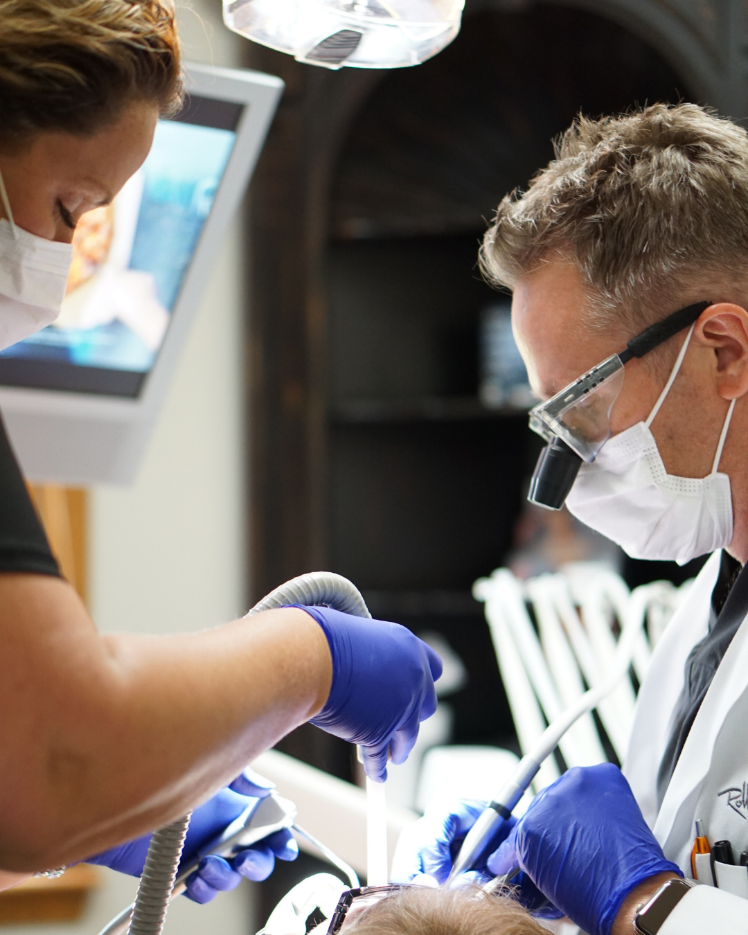 A dentist performs a procedure on a patient with a dental assistant holding a suction tool, both wearing masks and gloves, in a dental office.