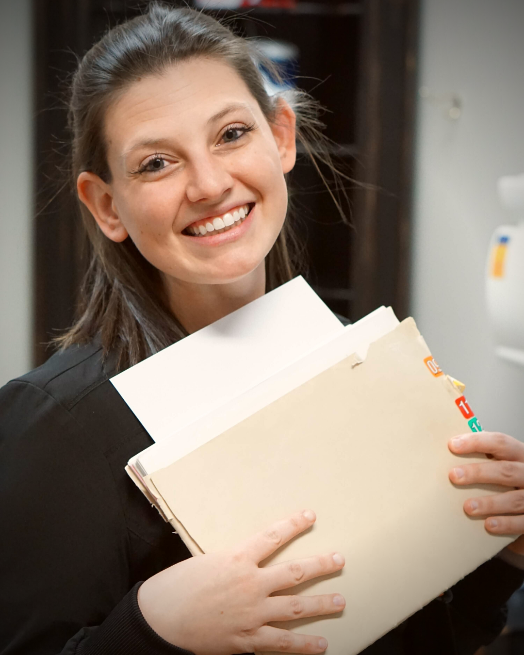 A woman with long brown hair smiling and holding a tan folder filled with papers.