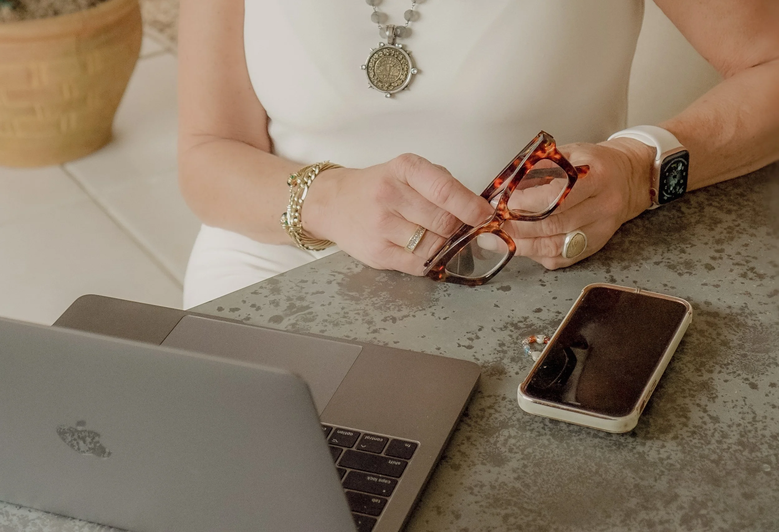 A person holding tortoiseshell eyeglasses above a gray countertop with a laptop, smartphone on the countertop, and wearing jewelry including a gold bracelet, ring, and a smartwatch.