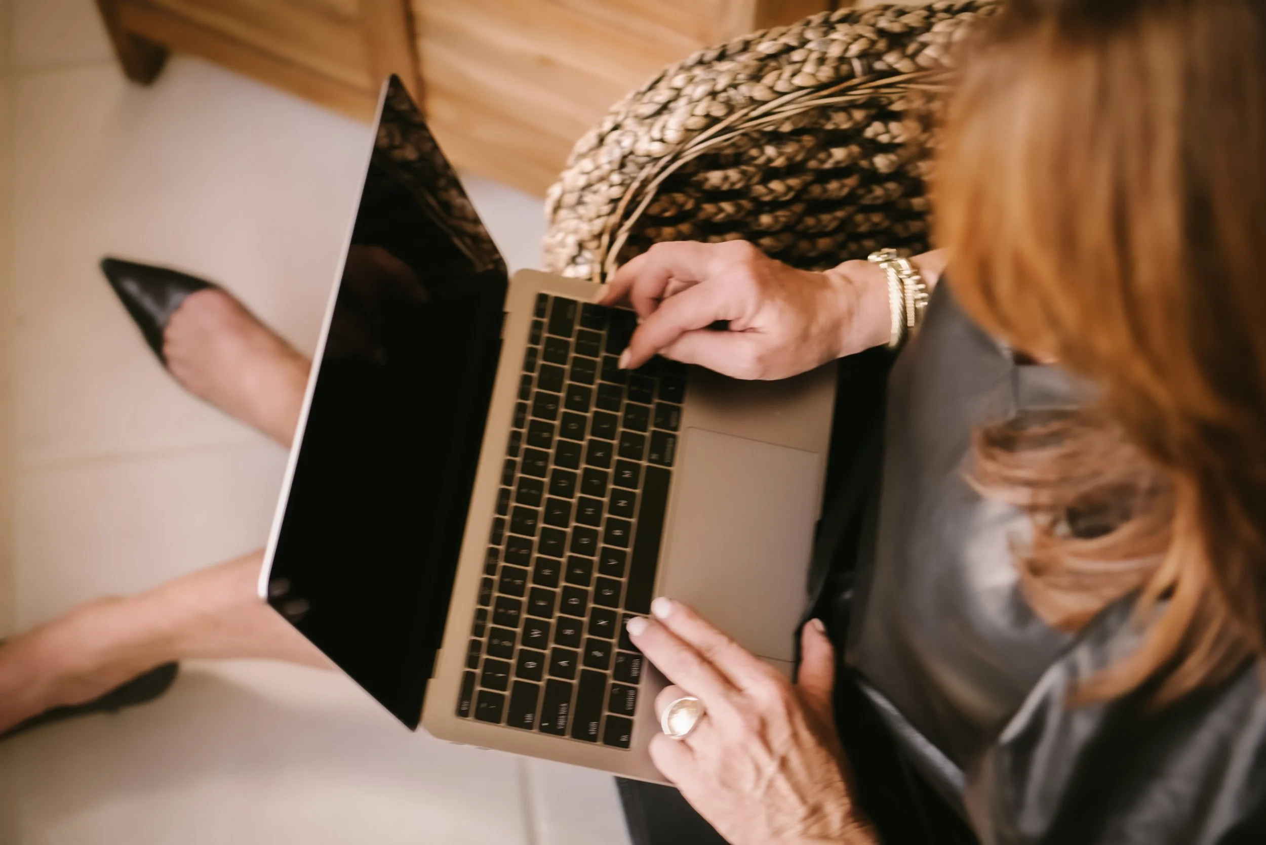 A woman with light skin and shoulder-length hair using a laptop on her lap, sitting on a light-colored chair with a person's legs visible in the background.