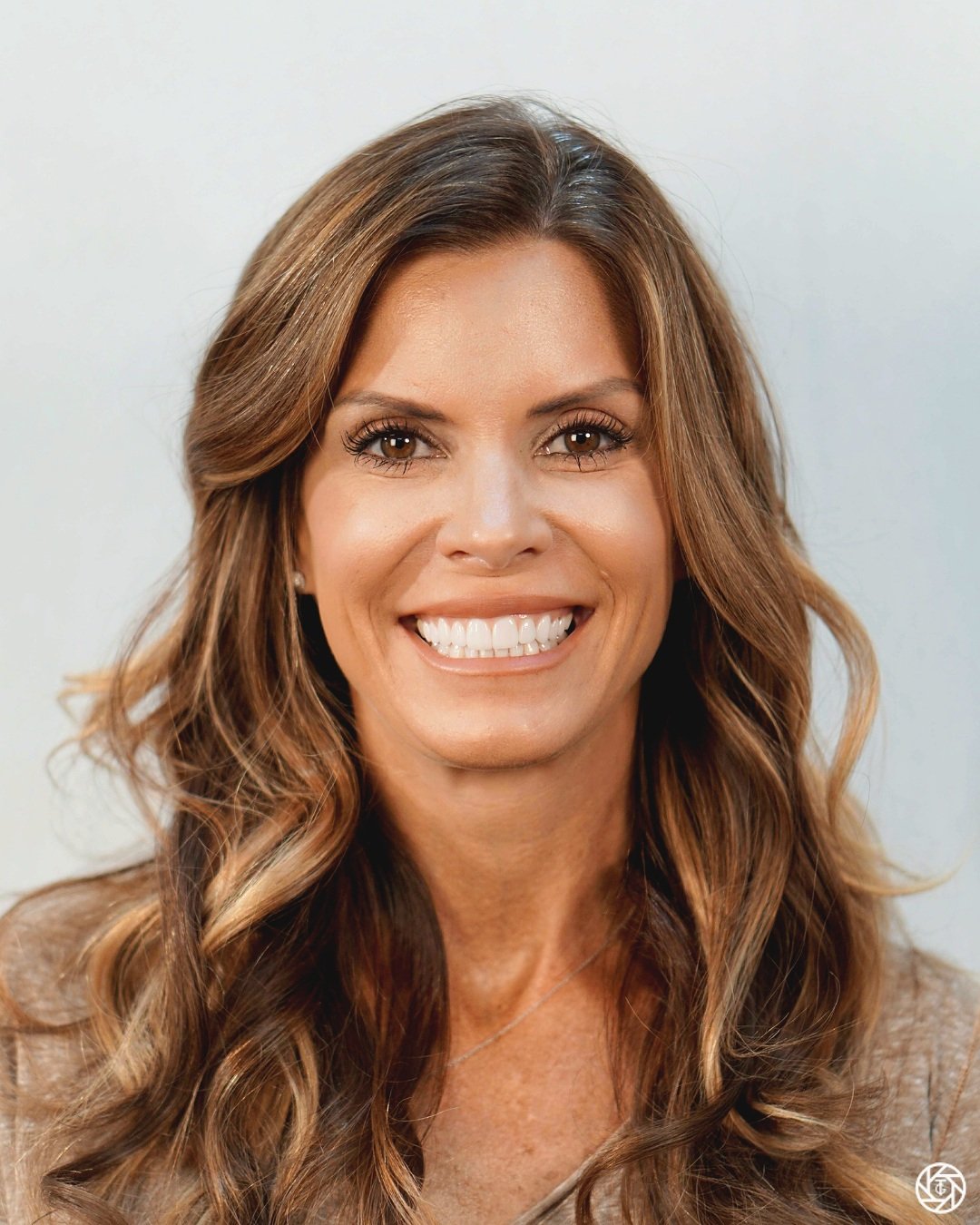 Close-up of a smiling woman with long, wavy brown hair and brown eyes, wearing light makeup and a beige top.