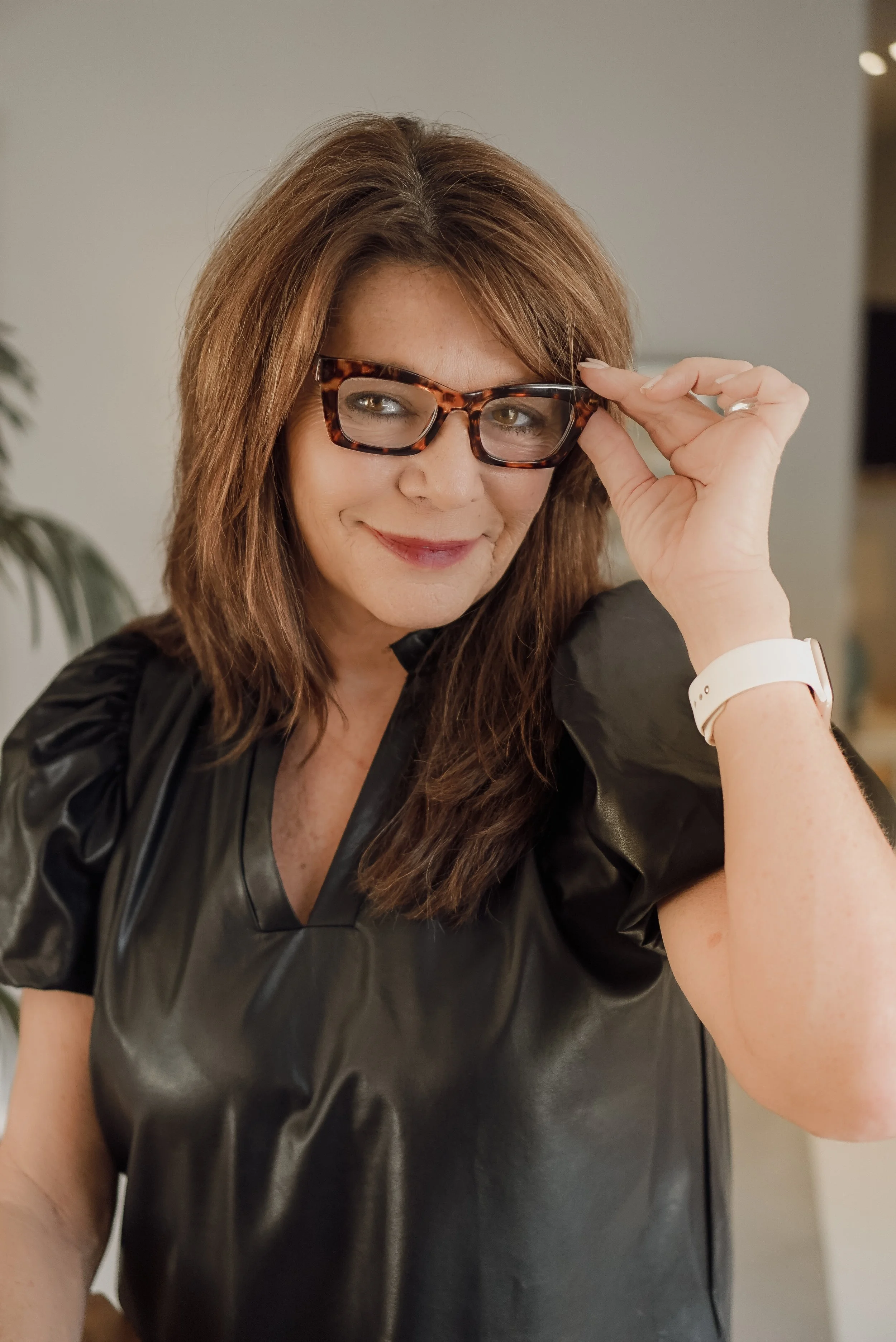 A woman with brown hair and glasses, smiling and adjusting her glasses with her right hand, wearing a black shiny top and a white wristwatch.