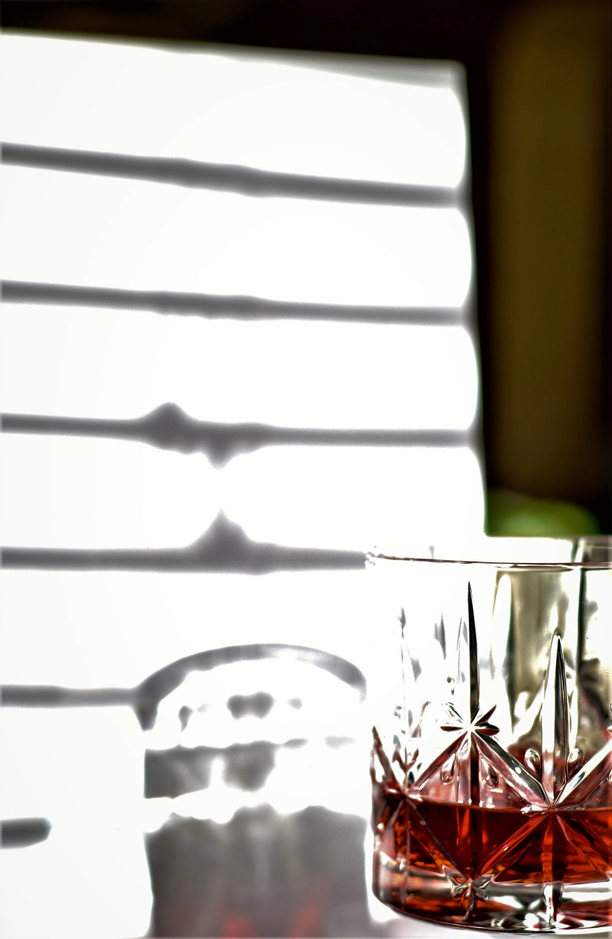  Crystal tumbler with Campari against shadows of venetian blinds. 