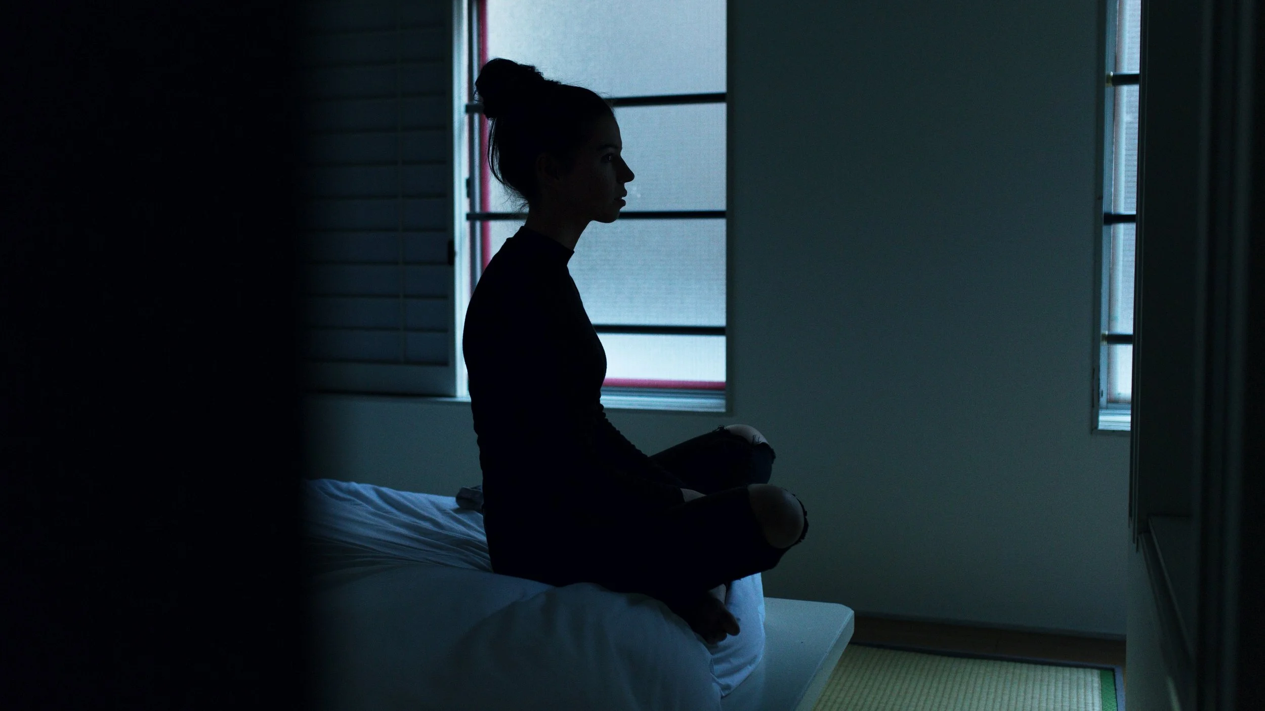 the silhouette of a woman sitting on the edge of her bed in a dark room. Her legs are crossed