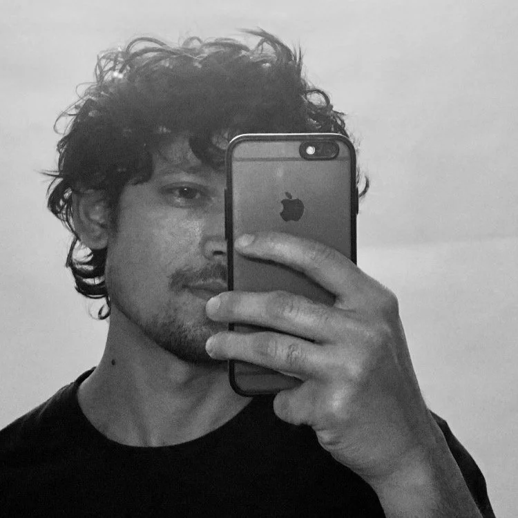 A young man with curly dark hair and facial hair takes a mirror selfie with an iPhone, partially covering his face, in black and white.
