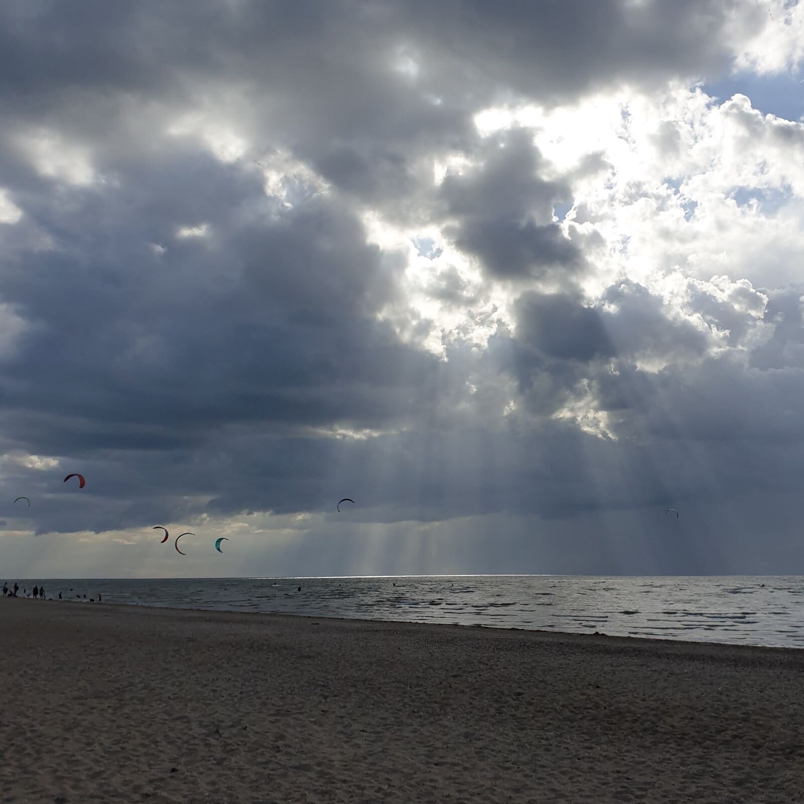 rays of sun breaking through grey clouds over a beach with kite surfers