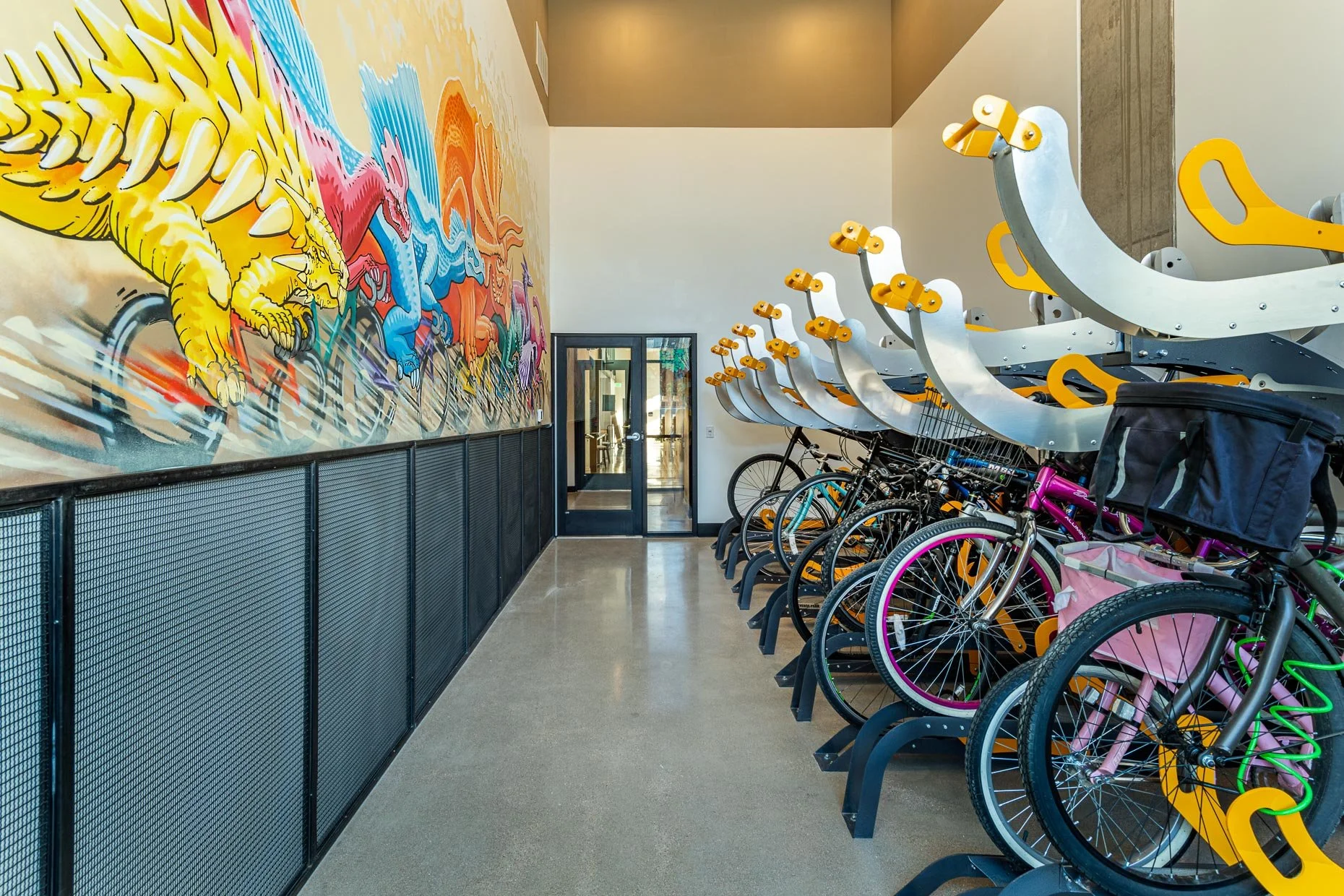 Bright indoor bike storage room with colorful dinosaur mural, polished concrete floor, and rows of bicycles mounted on sleek silver and yellow vertical racks along one wall.