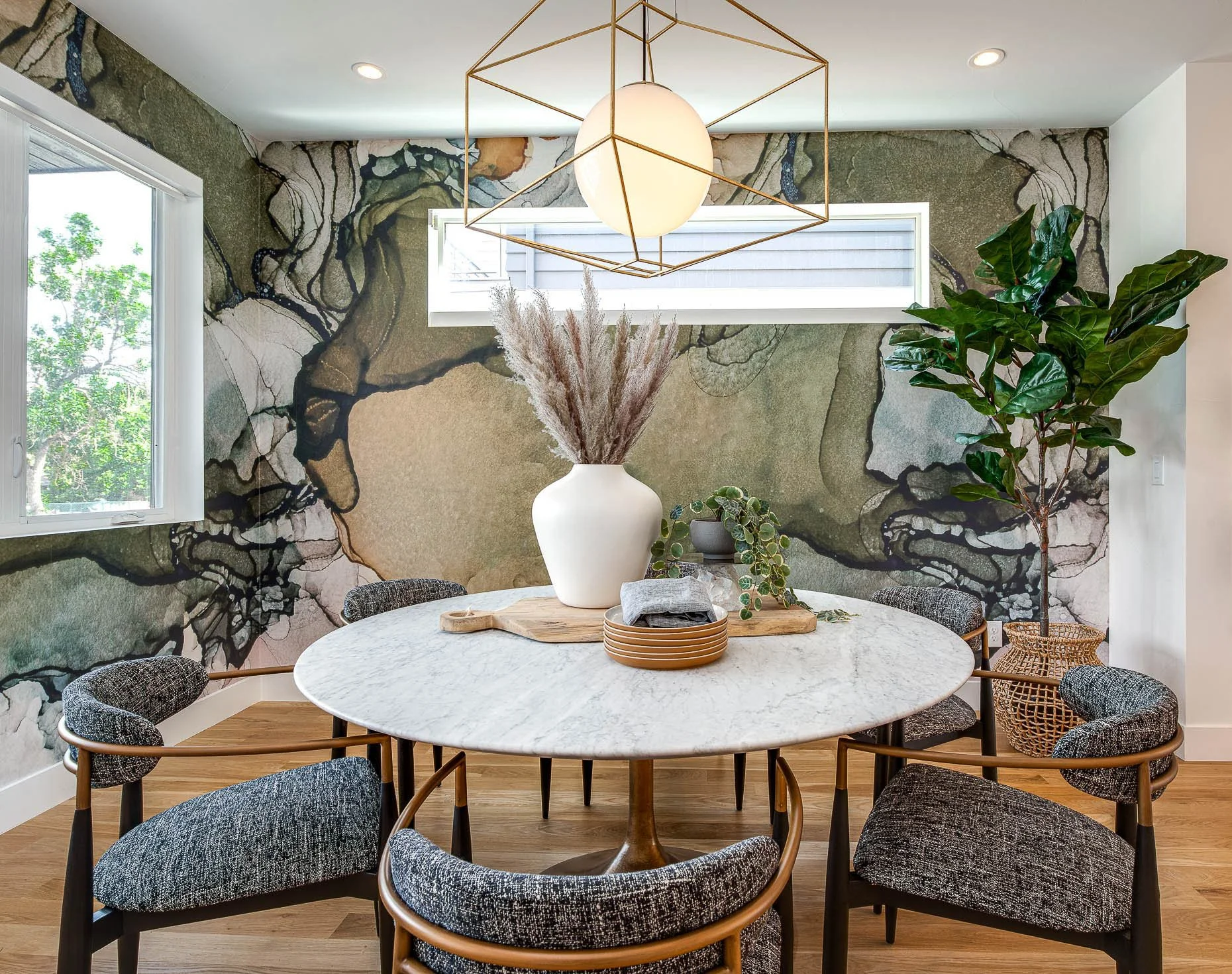 Stylish dining area featuring a round marble table with upholstered accent chairs, a geometric gold pendant light, and a bold abstract wallpaper in earthy tones.