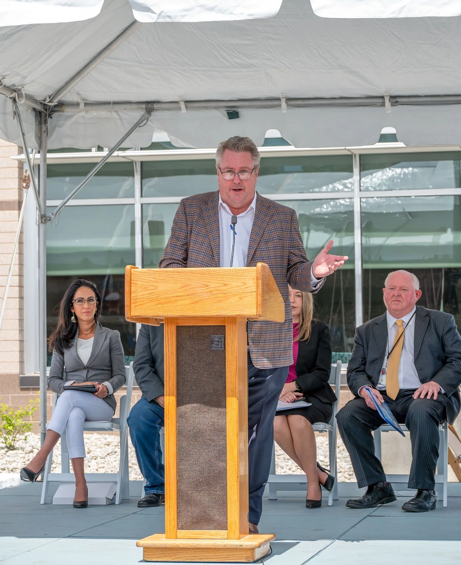 A man speaking at a podium during a formal outdoor event with three seated individuals behind him, under a large white tent.
