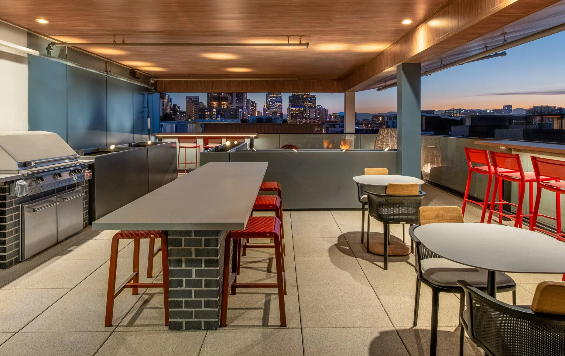 Covered rooftop lounge at AVA RiNo Apartments in Denver with outdoor kitchen, grill, modern seating, and bar-height tables overlooking the illuminated downtown skyline at dusk.