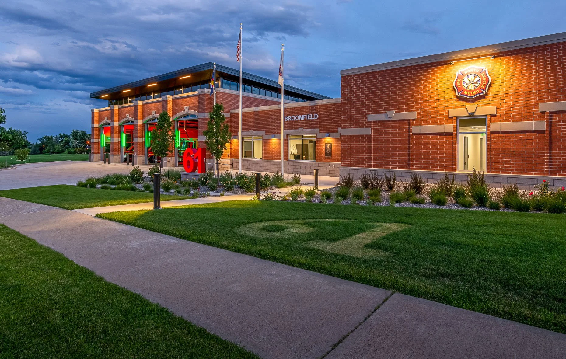 Dusk view of Fire Station 61 in Broomfield, Colorado, showing a modern red-brick firehouse with illuminated bays, landscaped grounds, three flagpoles, and a large glowing red “61” monument sign near the entrance.