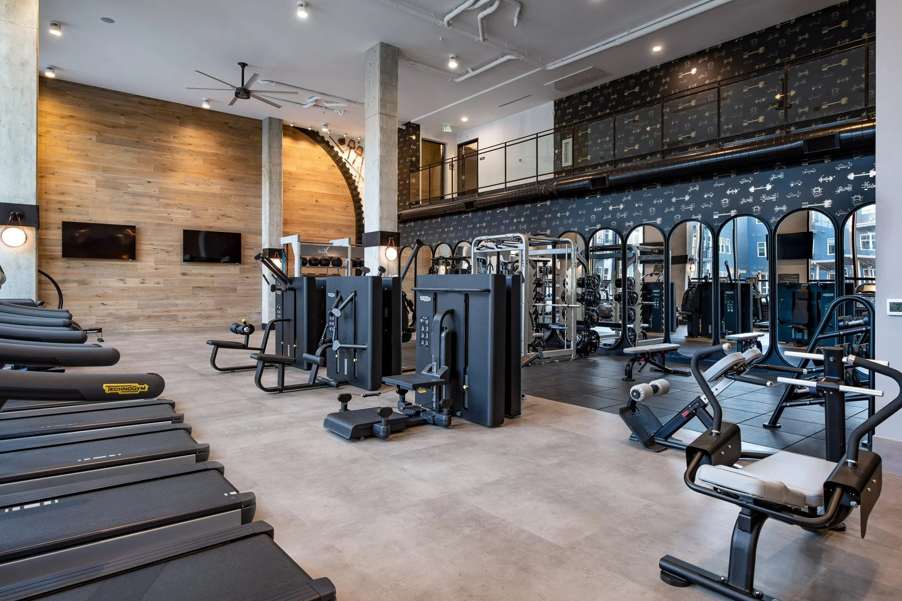 Spacious modern fitness center with treadmills, weight machines, and tall wood accent walls, featuring industrial concrete columns and a mezzanine level with black patterned wall and mirrors.
