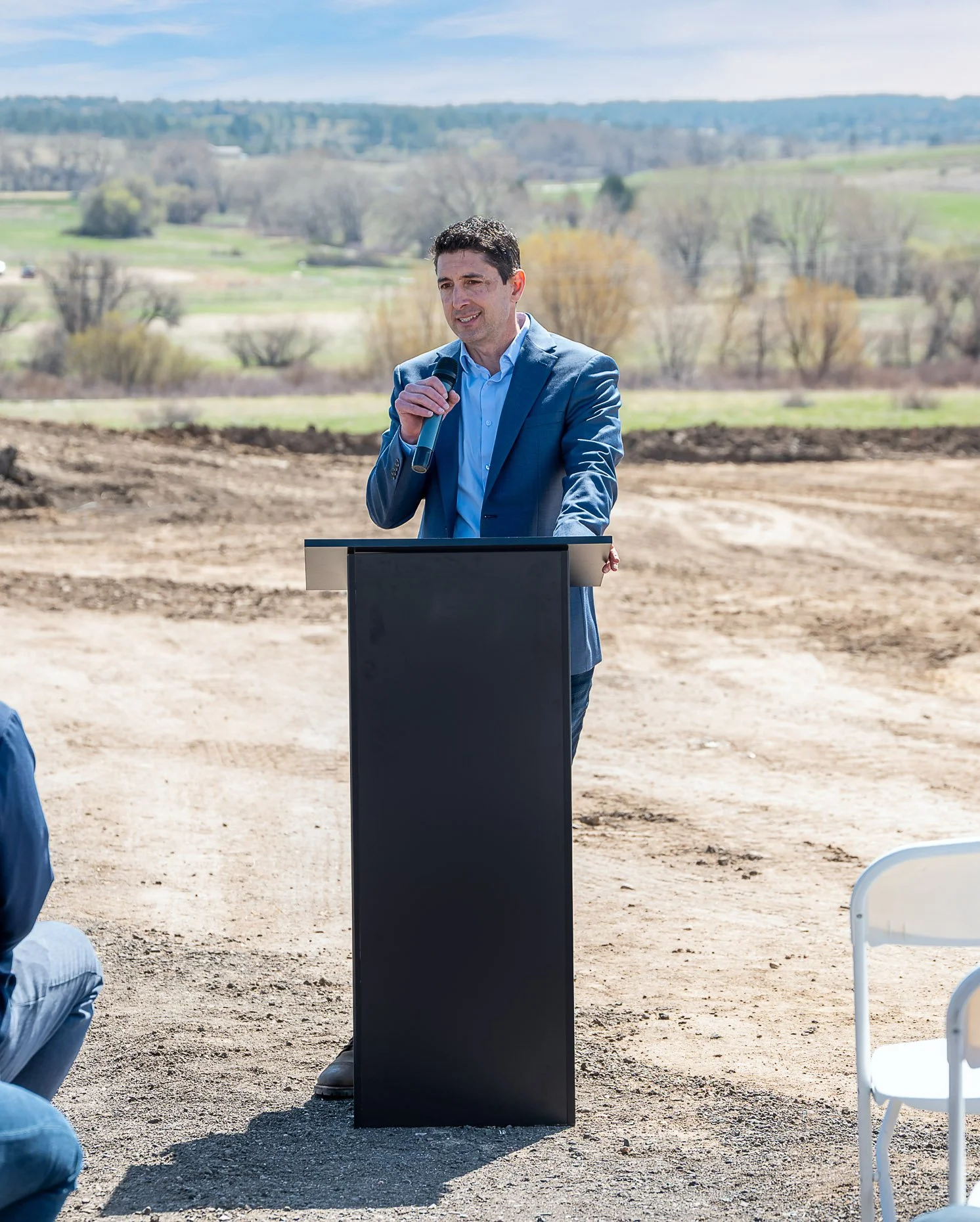 Man in a blue suit speaking at a podium outdoors with a microphone, in a rural area with trees and hills in the background.