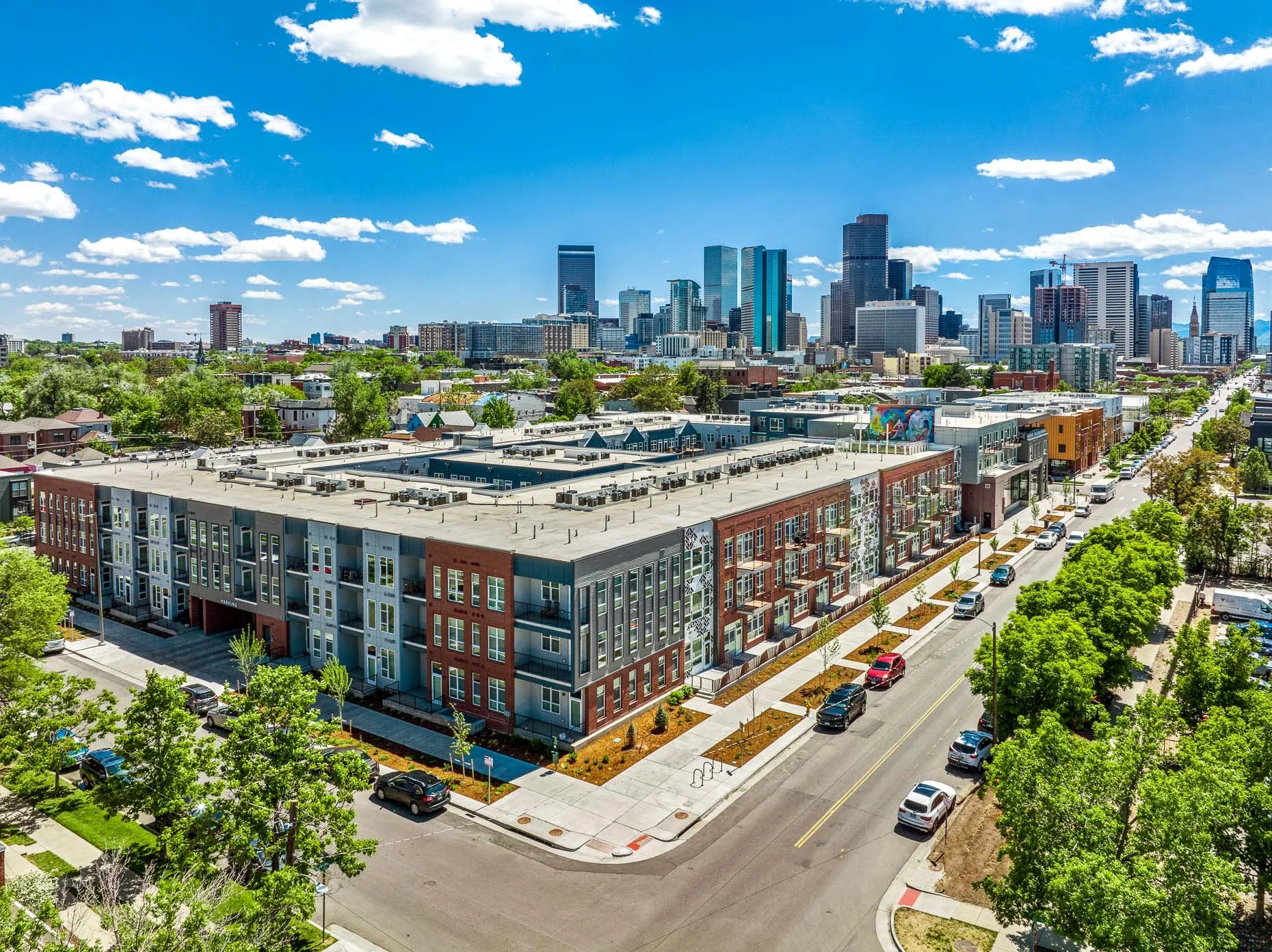 Aerial view of AVA RiNo Apartments in Denver with brick and modern facades, tree‑lined streets, and the downtown Denver skyline under a bright blue sky.