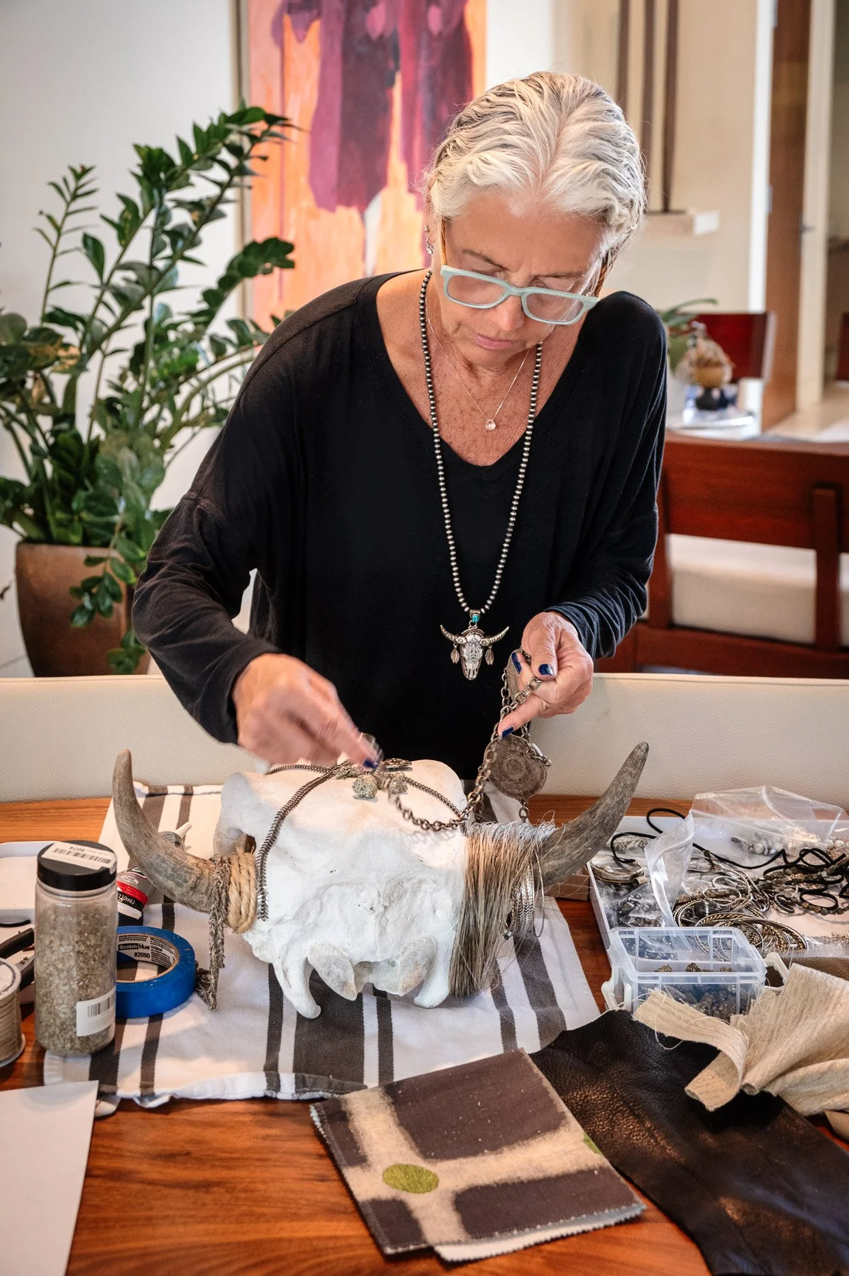woman with short gray hair, wearing glasses, necklaces, and a black top, is standing at a table covered with jewelry and accessories. She appears to be working with a large animal skull adorned with horns, which is also decorated with jewelry. The background features a piece of abstract art and a potted plant.