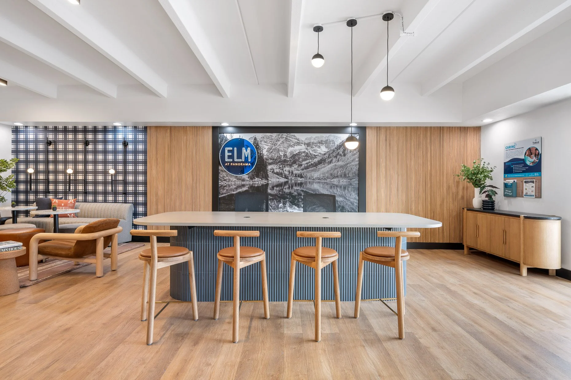 Modern leasing office at The Elm at Panorama featuring a long central island with four wood stools, warm wood flooring and wall panels, plaid accent seating area, and large “ELM at Panorama” mountain mural behind the desk.