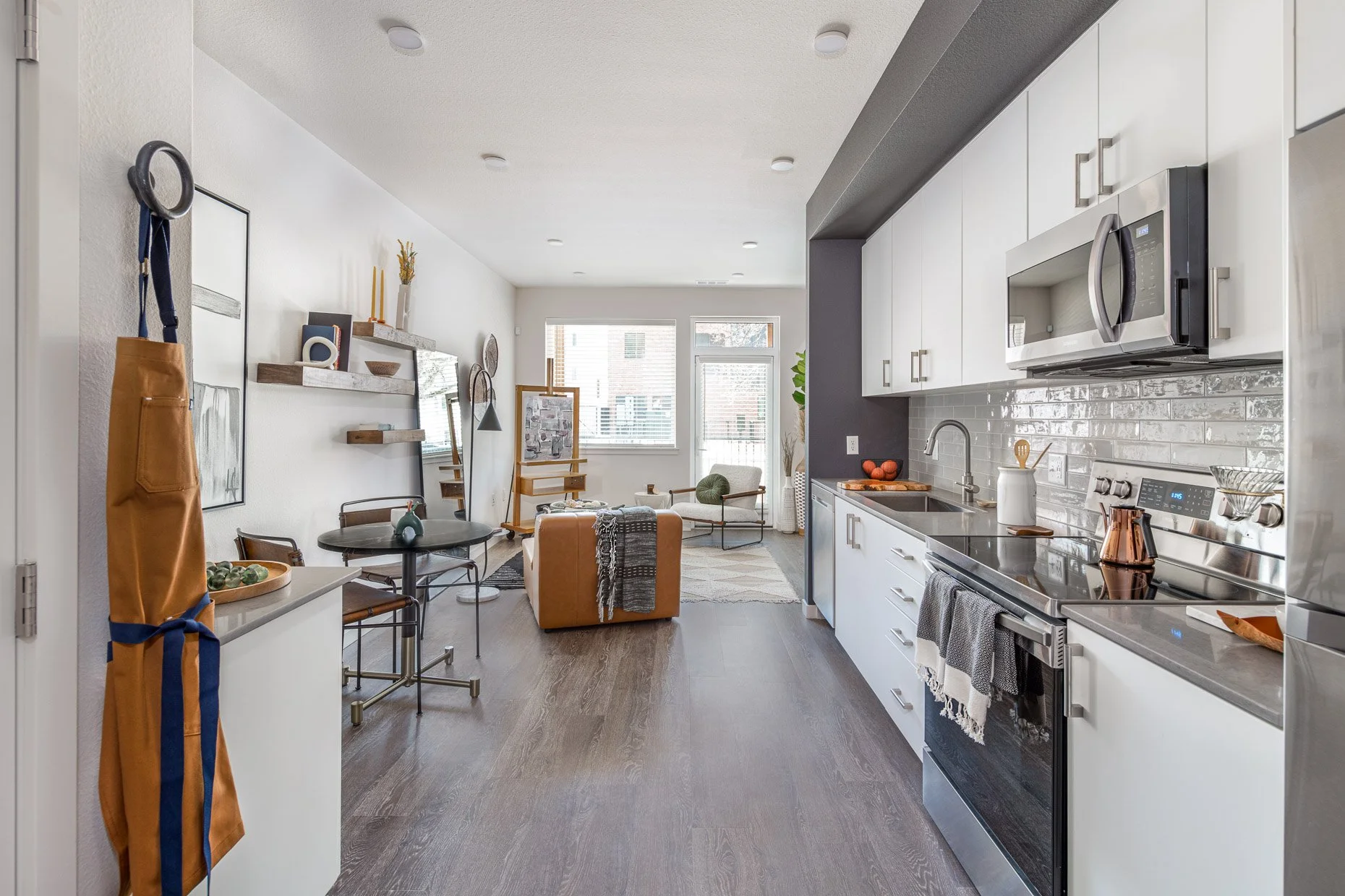 Open-concept AVA RiNo studio apartment interior featuring sleek white kitchen with stainless appliances, wood floors, and a bright living area with modern furnishings and large windows.