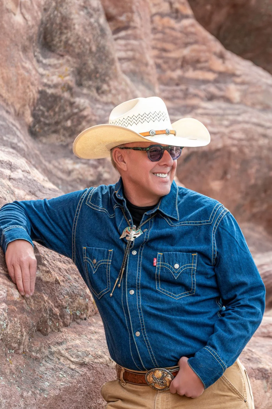 A man wearing a cowboy hat, sunglasses, and a denim shirt, smiling against a rocky desert landscape.