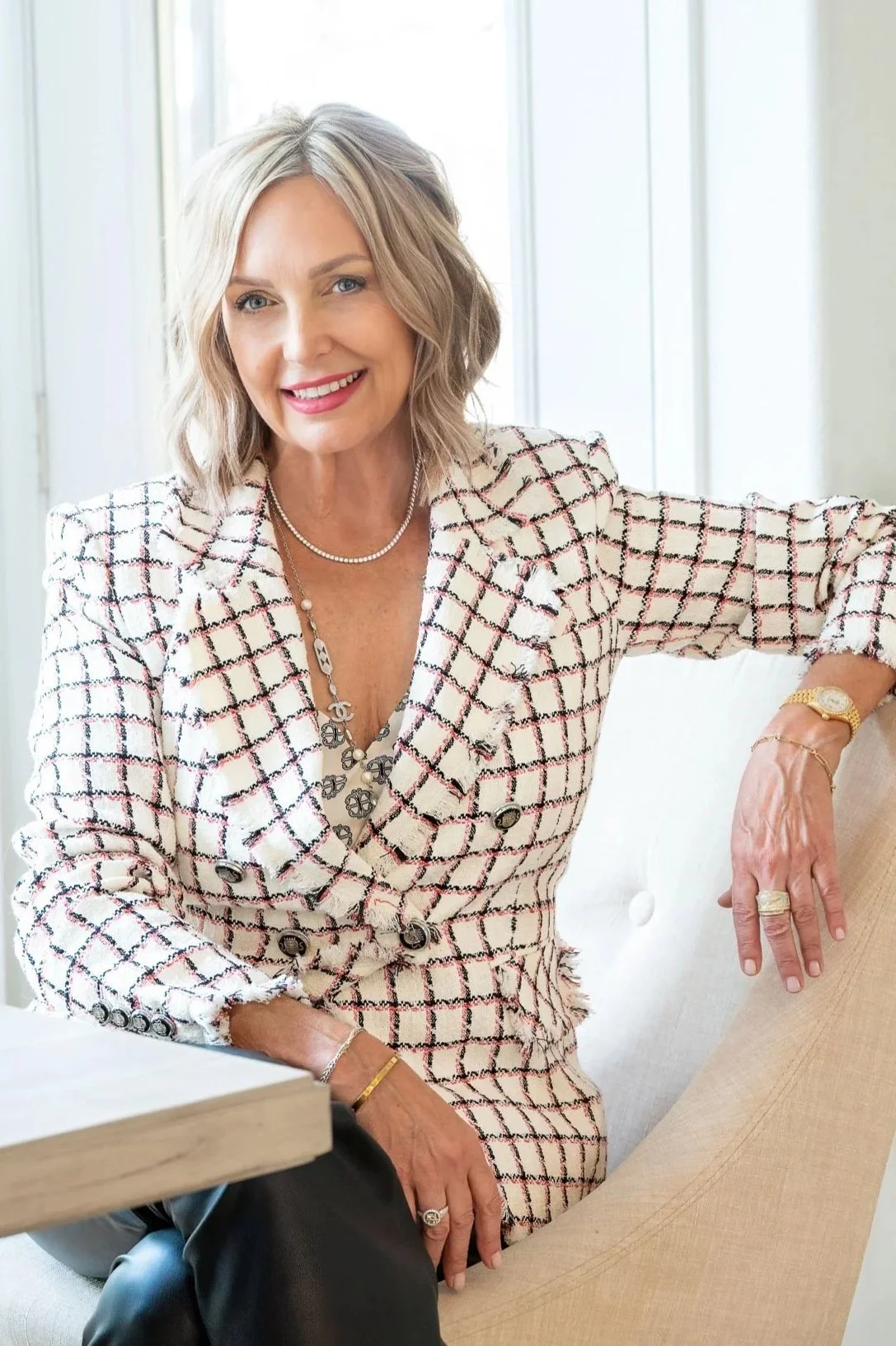 A smiling woman with shoulder-length blonde hair wearing a patterned blazer, sitting on a beige chair near a window.