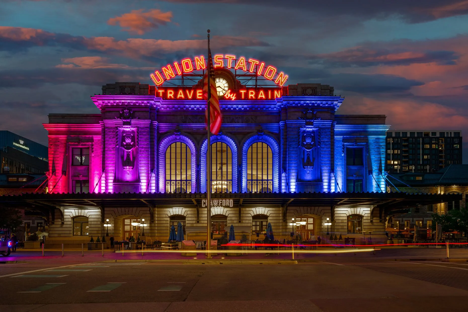 Dusk view of Denver’s Union Station with its “Travel by Train” sign glowing above the facade, illuminated in vibrant rainbow colors against a dramatic evening sky.