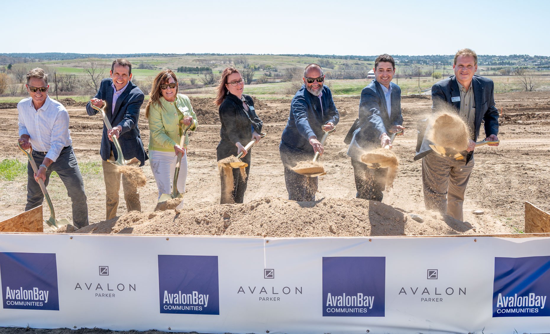 People in formal attire participate in a groundbreaking ceremony, each holding a shovel and tossing dirt on a construction site with open land in the background. A banner with logos for AvalonBay Communities and Avalon Parker is displayed at the front.