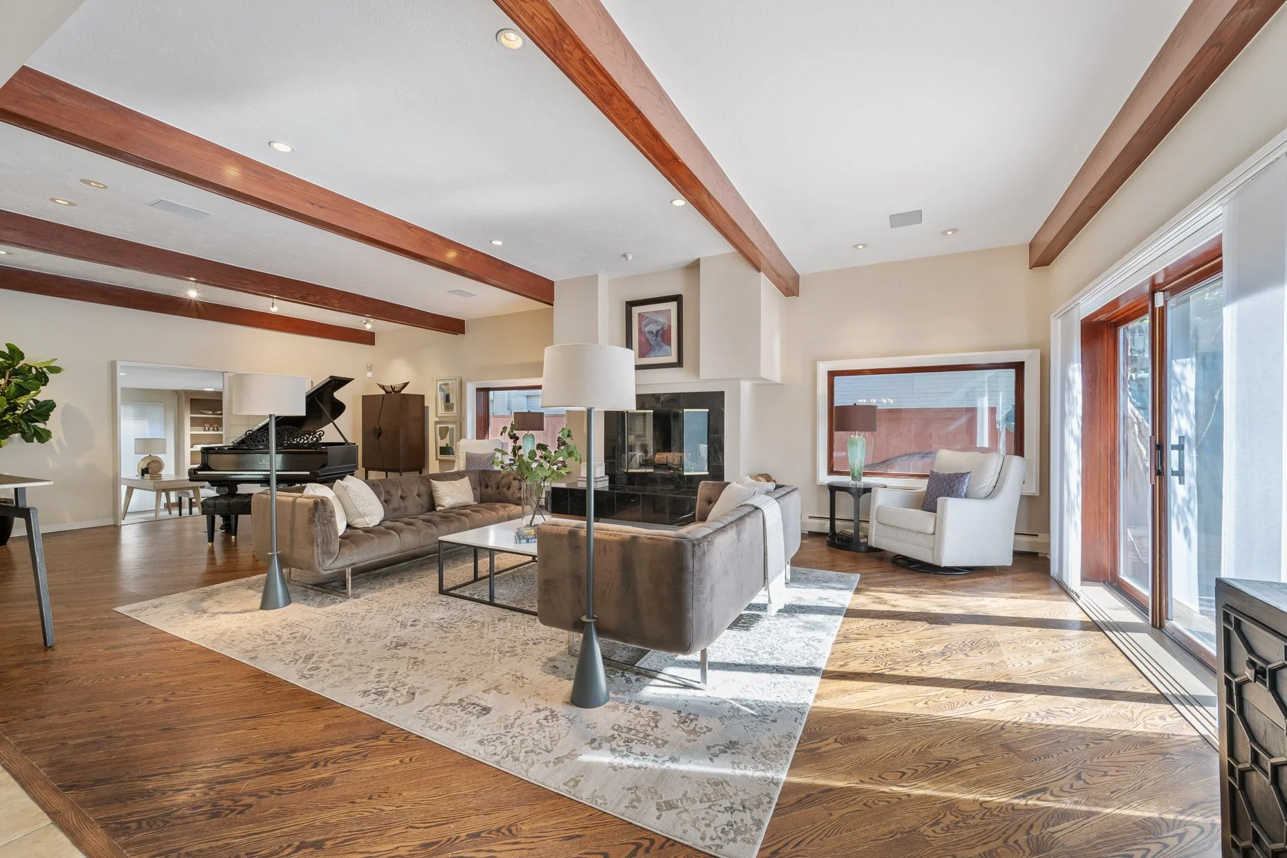 Spacious living room with hardwood floors, white walls, large windows with wooden frames, and a fireplace. Furniture includes two brown sofas, a white armchair, a black piano, and side tables with lamps. Natural light fills the room.