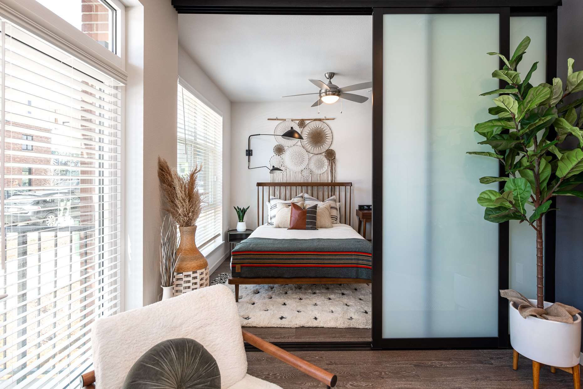 Stylish AVA RiNo studio bedroom with modern wood bed, layered neutral bedding, wall art, sliding frosted glass door, and large windows bringing in natural light.