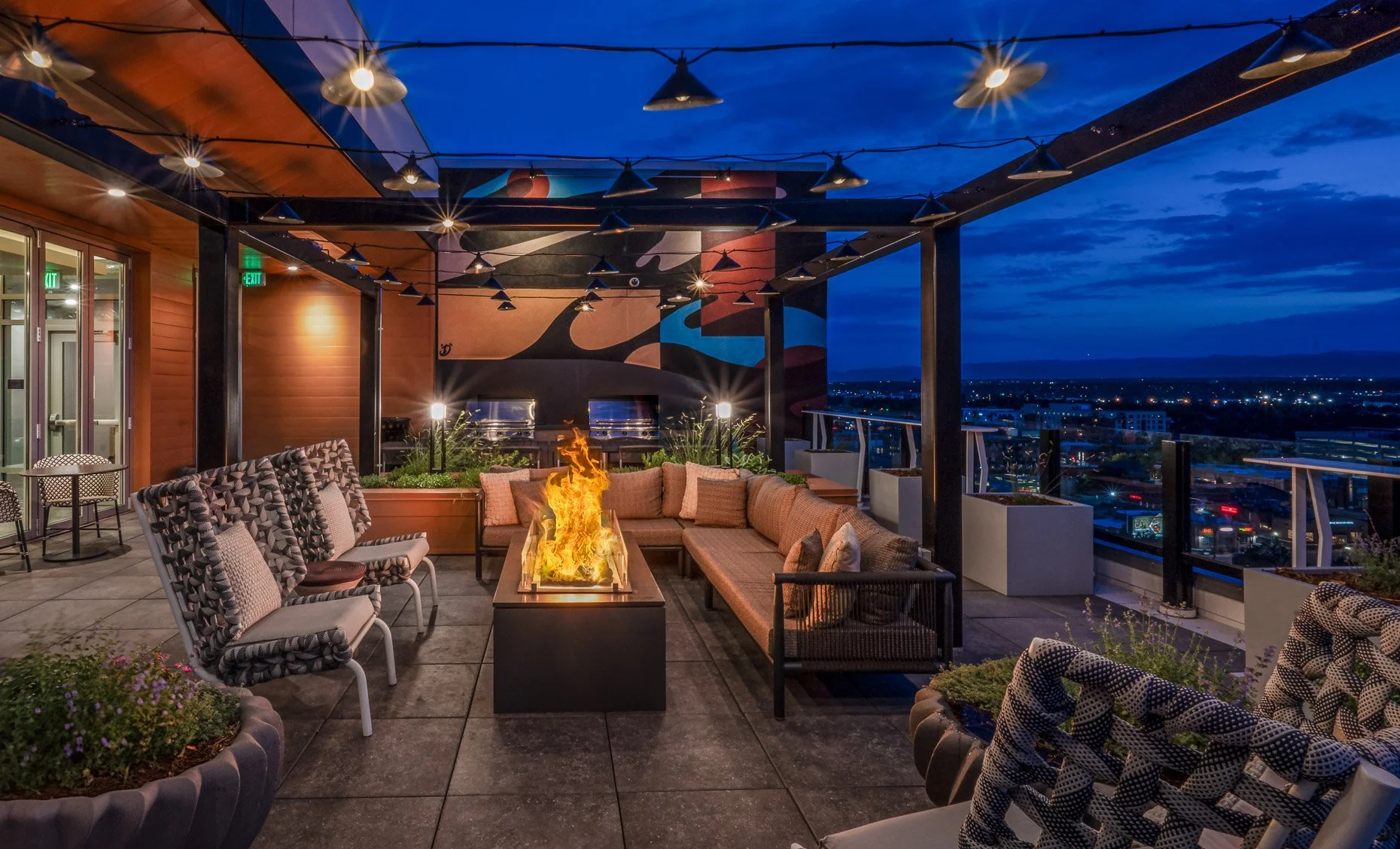 Rooftop lounge at AvalonBay Governor’s Park Apartments in Denver at dusk featuring a modern fire pit, cozy sectional seating, string lights, colorful mural wall, and expansive city views under a deep blue sky.