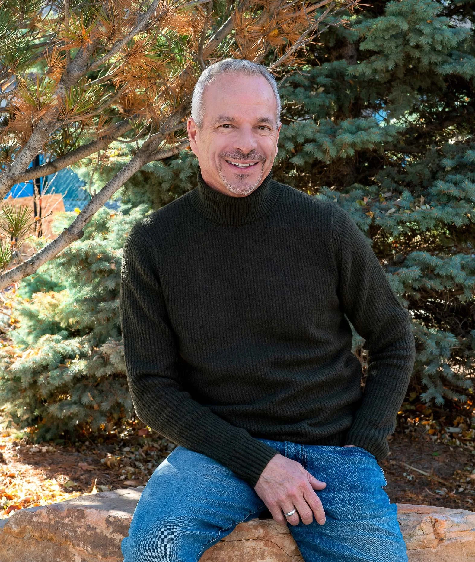 A smiling middle-aged man with short gray hair, beard, and mustache, wearing a black turtleneck sweater and blue jeans, sitting on a large rock outdoors surrounded by green and brown pine trees.