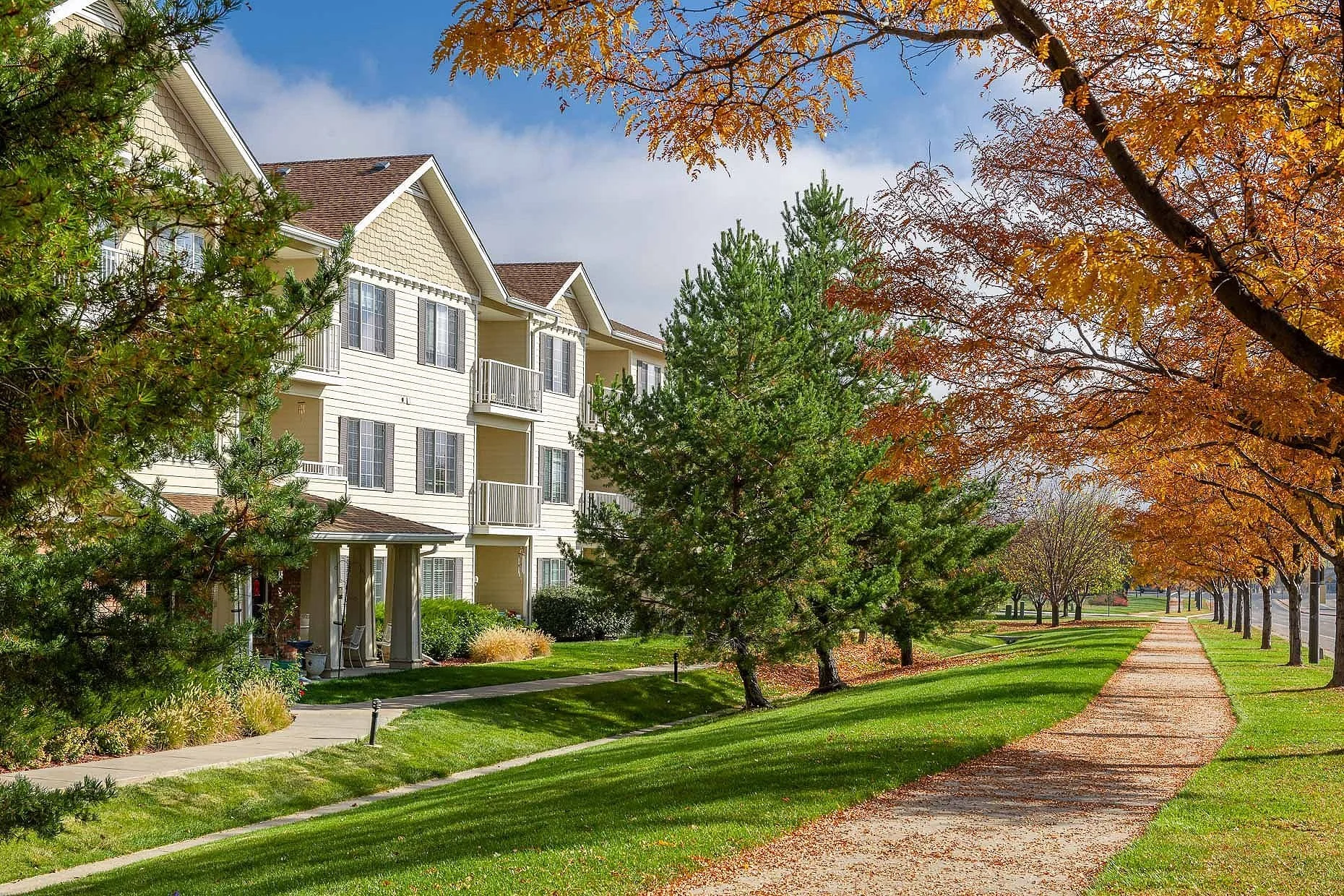 Charming 55+ apartment complex surrounded by lush greenery and a scenic walking path lined with vibrant autumn trees. The three-story beige building features private balconies and a welcoming front porch, creating a serene community setting.