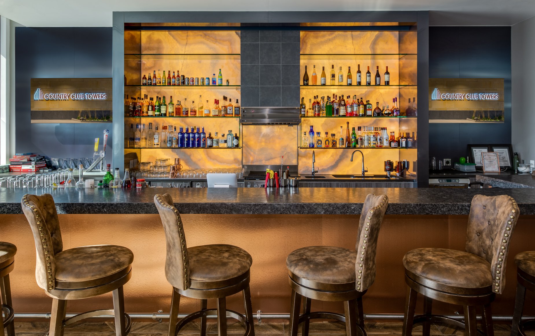 Elegant community bar at Broe Country Club Estates Condos featuring a long stone countertop with upholstered barstools, illuminated backlit onyx shelves stocked with bottles, and sleek modern signage on the surrounding walls.