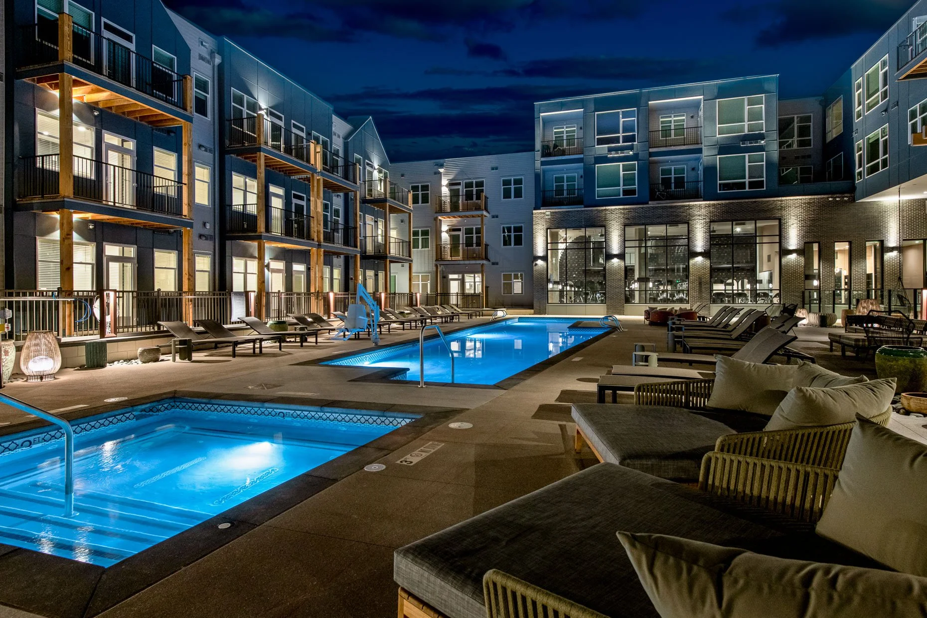 Nighttime view of AVA RiNo courtyard pool deck in Denver featuring glowing pool and hot tub, lounge chairs, and modern apartment buildings lit with warm exterior lighting.