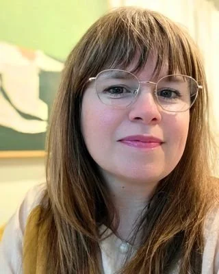 Close-up of a therapist, Amanda, with long light brown hair, wearing glasses, smiling in front of a green wall with a canvas painting in the background.