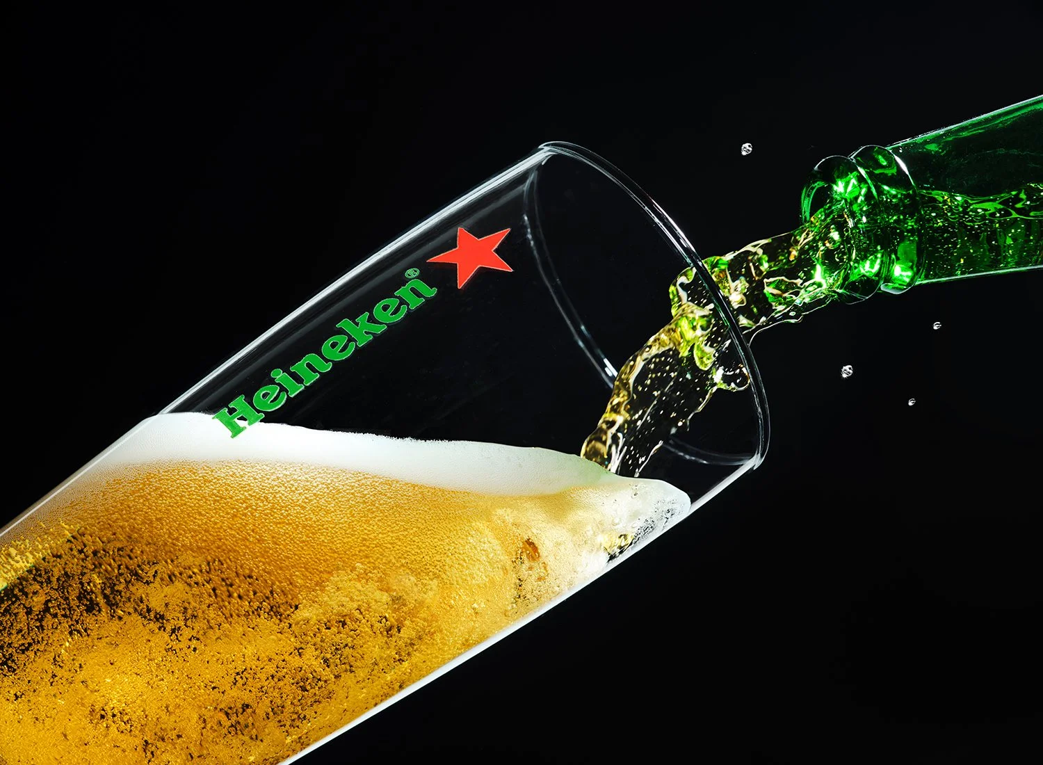 Action still life photograph of beer pouring from a Heineken bottle into a branded glass, shot in studio by Takamasa Ota, New York City.