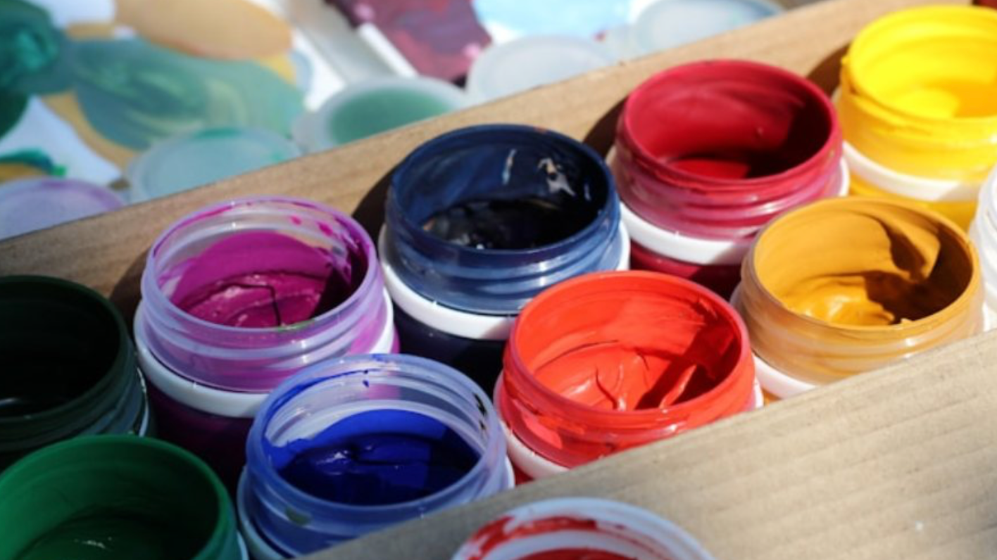 Open containers of various colors of paint, including purple, blue, red, yellow, black, and green, arranged in rows on a wooden surface.
