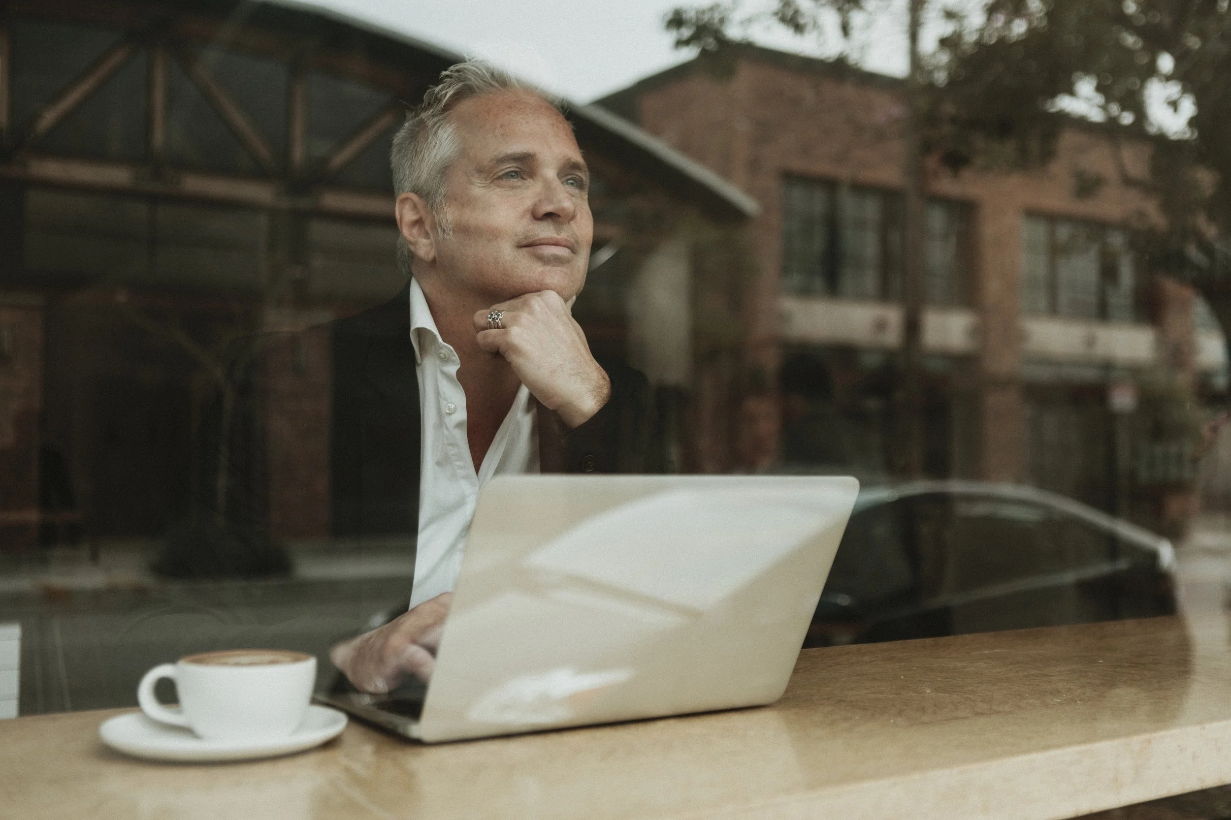 A man sitting at a coffee shop table with a laptop and a cup of coffee, looking thoughtfully out the window.