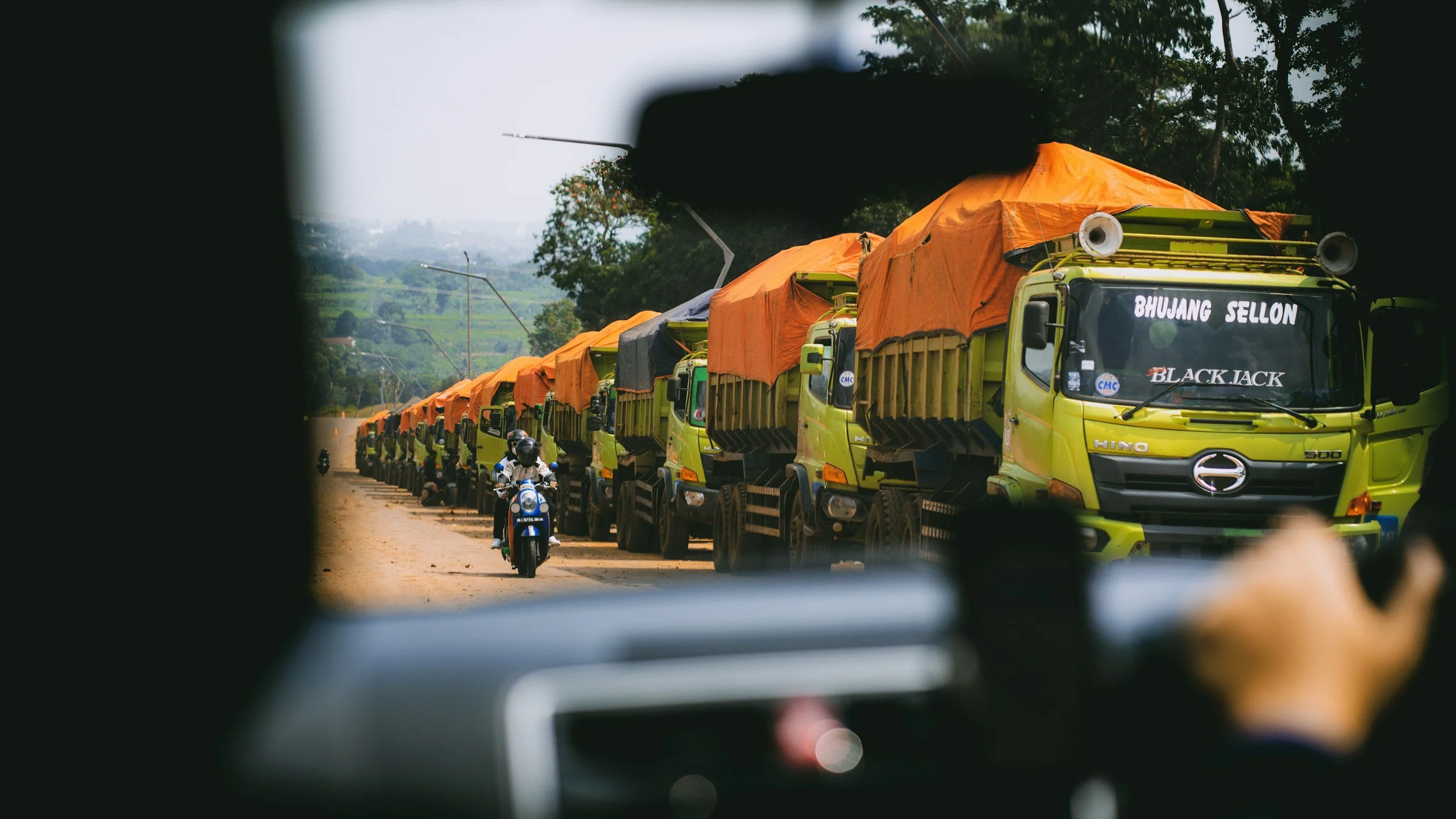 Line of green trucks with orange tarps on the road, a motorbike and rider in front, and trees and hills in the background, viewed through a car window.