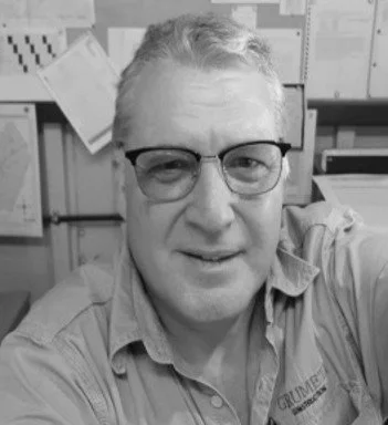 A man wearing glasses and a collared shirt, smiling at the camera in an office or storage room environment.