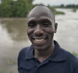A smiling man outdoors near a river or large body of water with trees in the background.