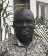 Portrait of a man with dark skin in front of greenery, wearing a patterned shirt and smiling.
