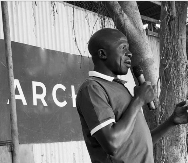 A man speaking into a microphone outdoors, standing beside a tree during daytime.