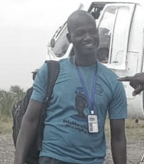 A man smiling outdoors, wearing a blue shirt, carrying a backpack, and a lanyard with an ID badge. There is an aircraft in the background.