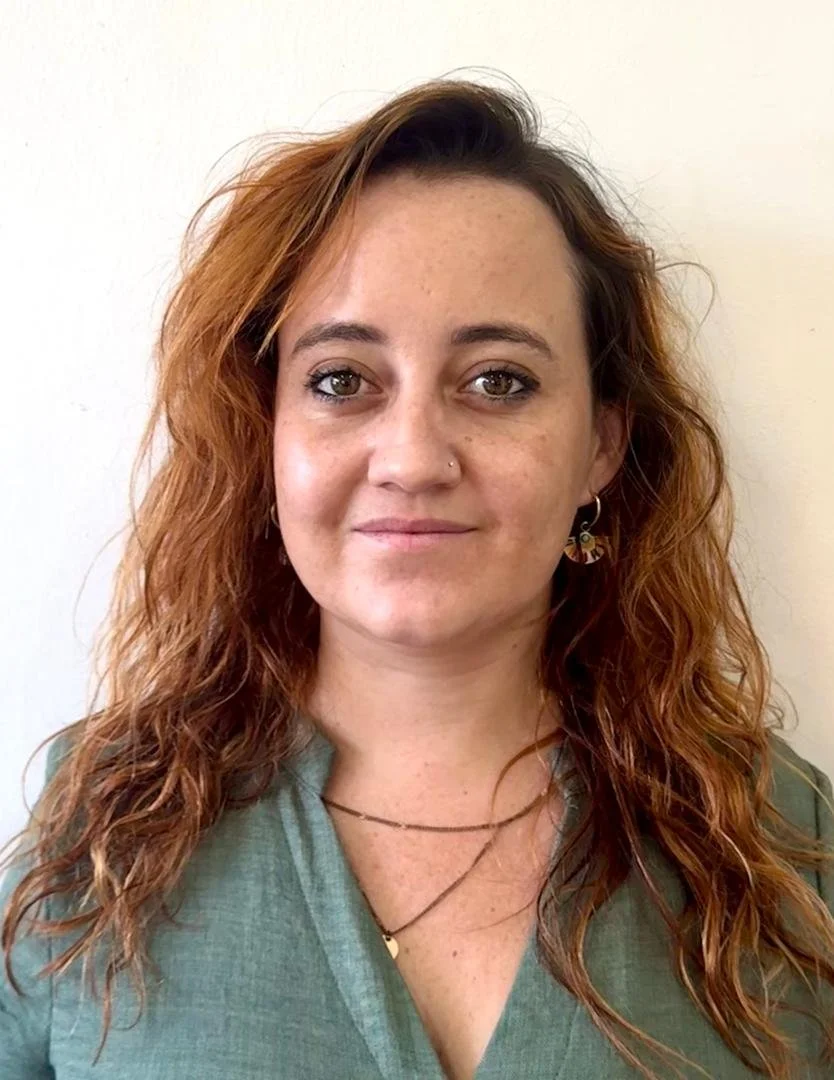 A young woman with reddish, curly hair, light skin, and hazel eyes, wearing a green top, earrings, and layered necklaces, standing against a plain white background.