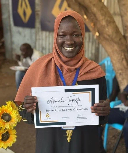 Woman wearing a brown headscarf holding a certificate with a bouquet of sunflowers nearby, outdoors at an event.