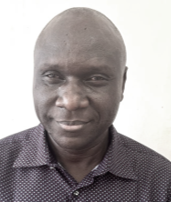 Portrait of a man with dark skin, bald head, wearing a dark patterned shirt, smiling against a plain white background.