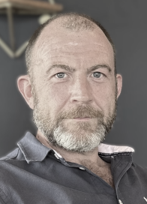 A man with a beard and short hair, wearing a dark collared shirt with white detail, looking directly at the camera, against a blurred grey background.