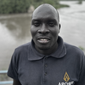 Portrait of a man outdoors near a body of water, wearing a black polo shirt with a logo on it.