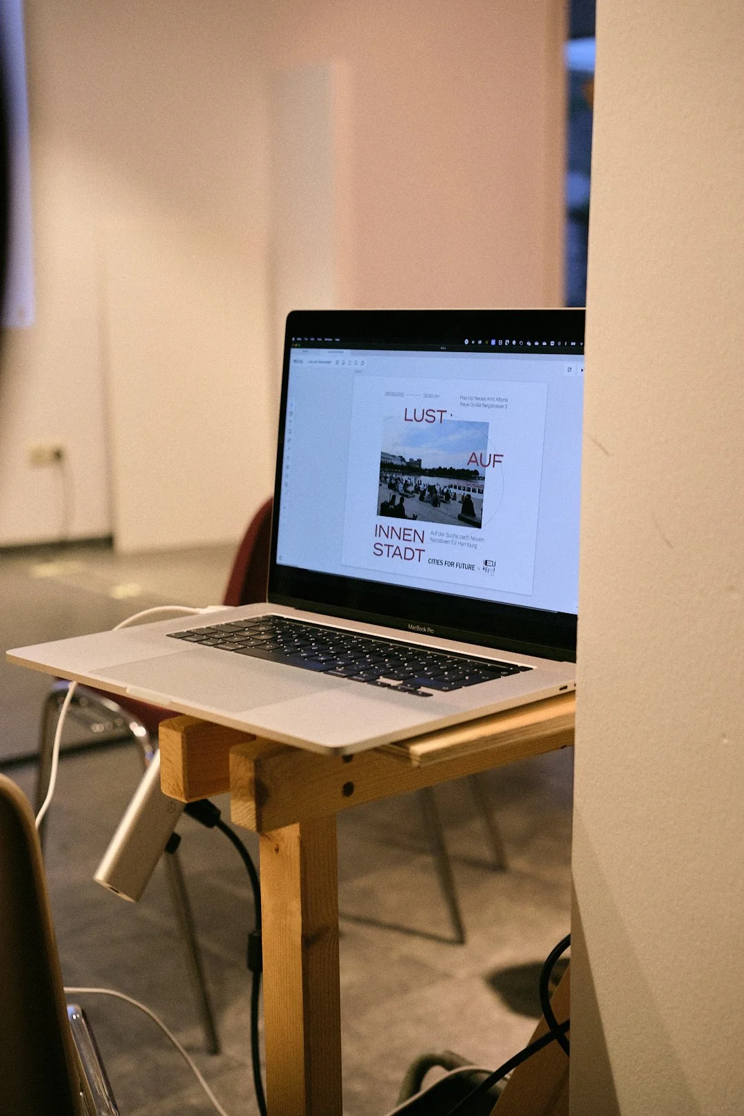 Ein Laptop auf einem Holztisch, geöffnet mit einem Dokument auf dem Bildschirm, in einem Raum mit weißen Wänden und Stühlen im Hintergrund.