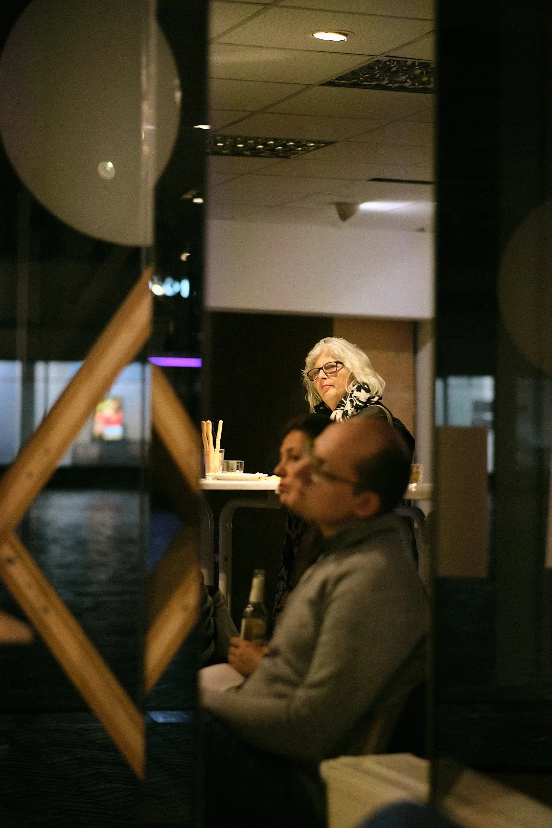Menschen in einem Restaurant, im Vordergrund ein Mann mit Brille, im Hintergrund eine ältere Frau mit Graublonden Haaren und Brille, die an einer Bar steht.